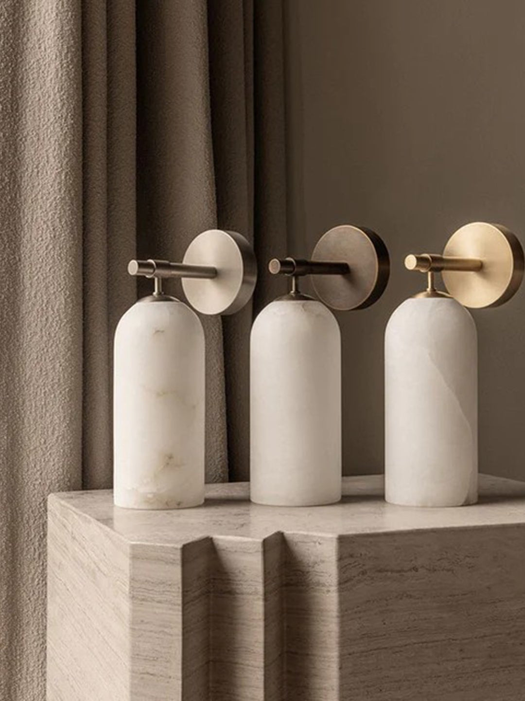 Three Clérachic Pillar wall lights lined up on a stone plinth in brass and bronze finishes.