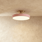 Clérachic ceiling light in dusty pink with a brass stem, its round drum glowing warm white against a plaster ceiling.