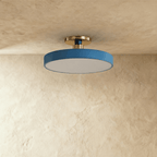 Clérachic ceiling light in blue with a brass stem, its round drum diffuser glowing warm against the ceiling.