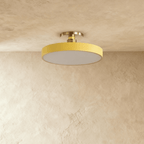 Clérachic ceiling light in yellow, its round drum diffuser glowing warm white beneath a plaster ceiling.