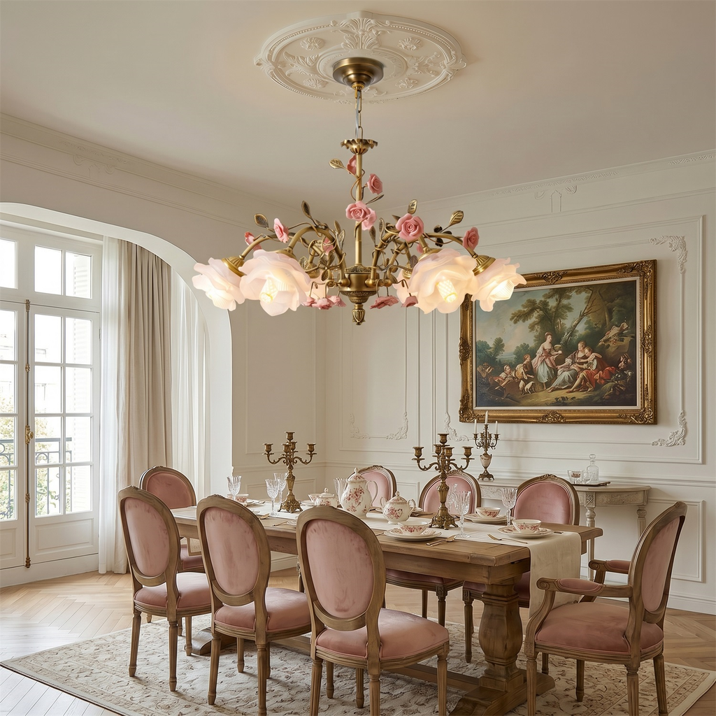 A lit Clérachic rose chandelier hung from a plaster ceiling rose over a wooden dining table with pink velvet chairs and a gilt-framed painting.