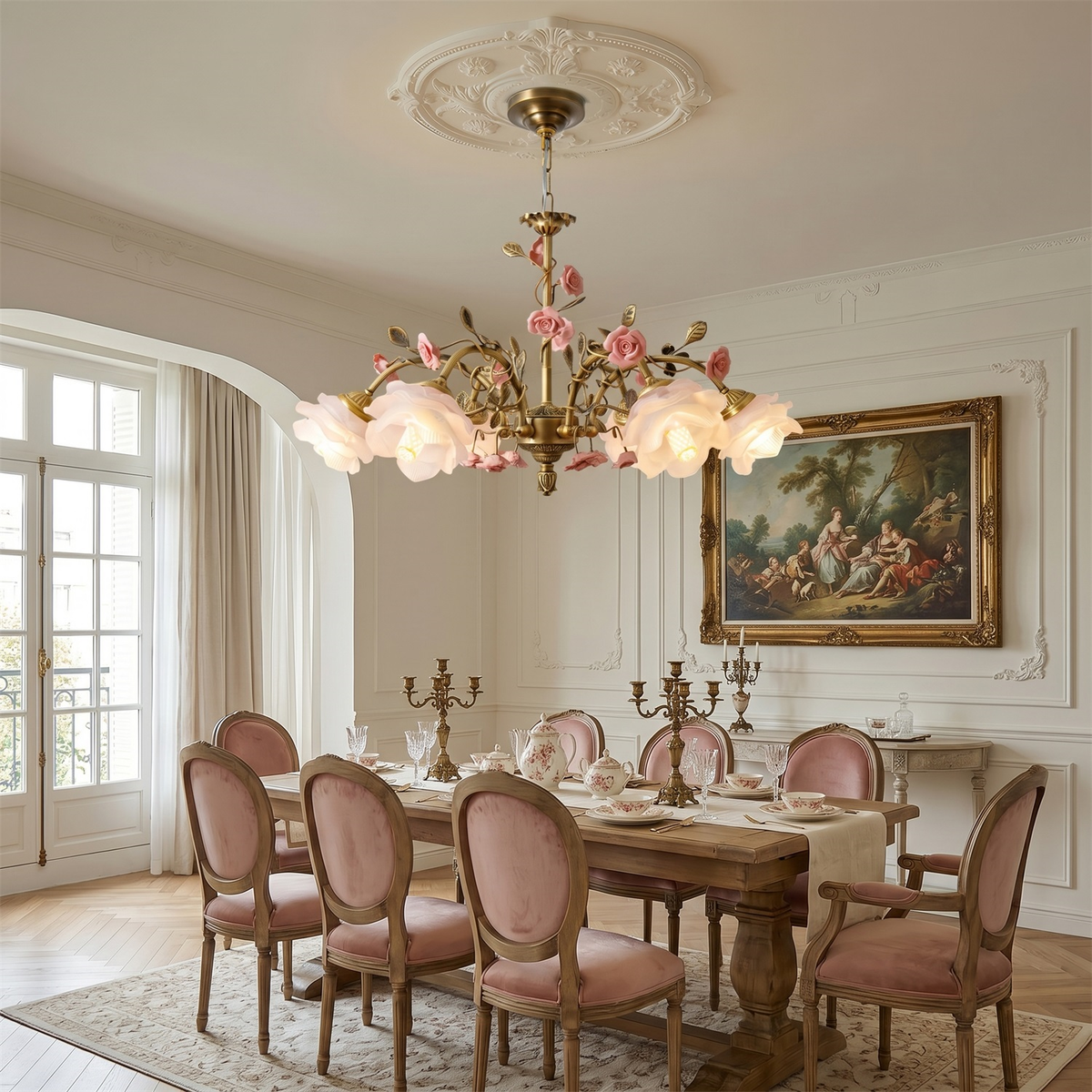 A lit Clérachic rose chandelier hung from a plaster ceiling rose over a wooden dining table with pink velvet chairs and a gilt-framed painting.