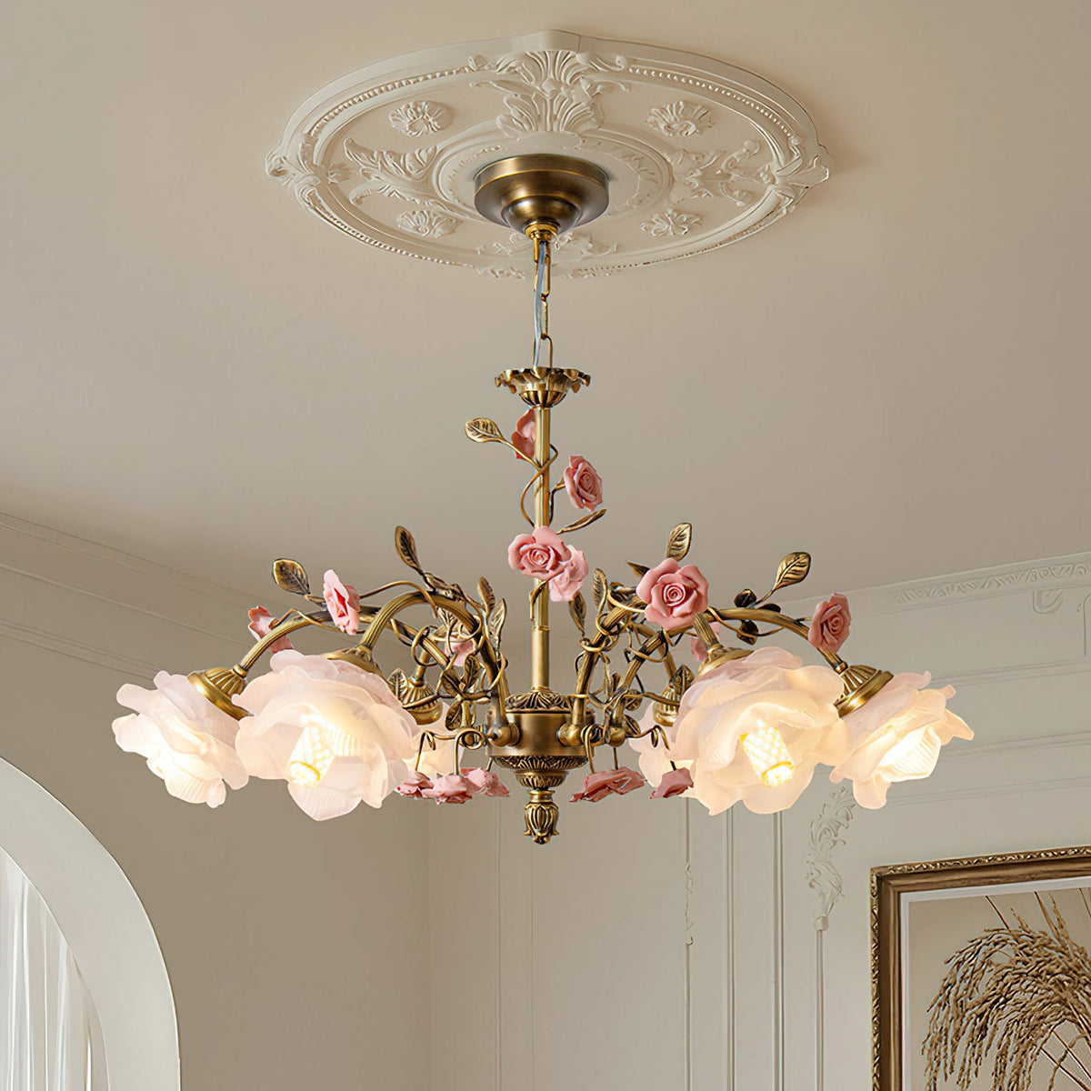 Close view of this Clérachic chandelier hanging from a decorative white ceiling rose, with lit frosted rose shades and ceramic flowers on gold branches.