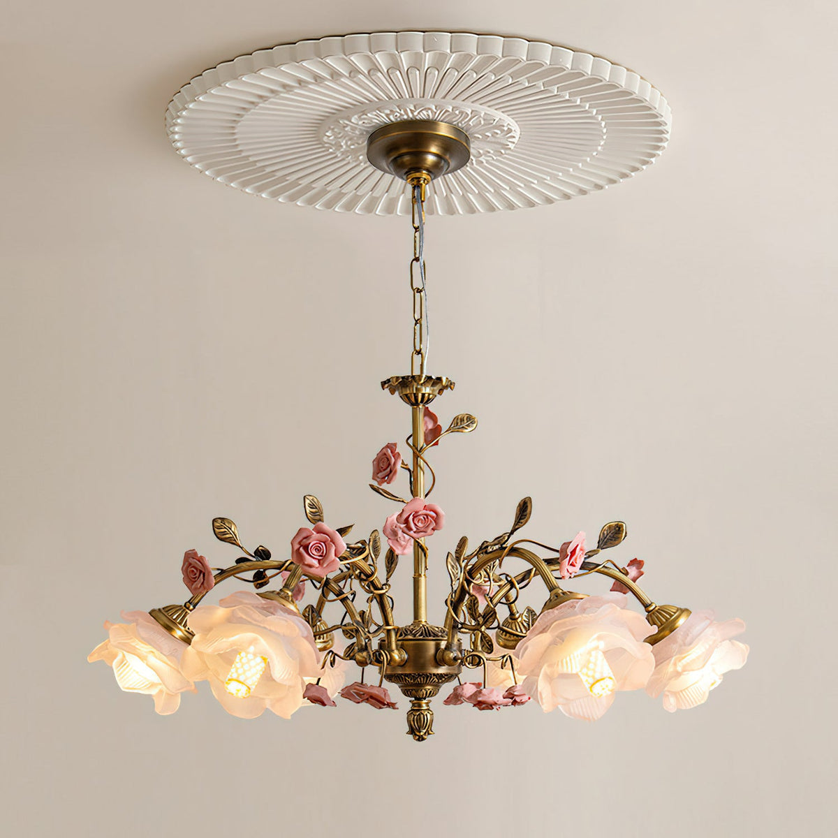 Clérachic chandelier lit beneath a round fluted ceiling rose on a pale wall, its antique gold chain and arms trailing small pink roses.