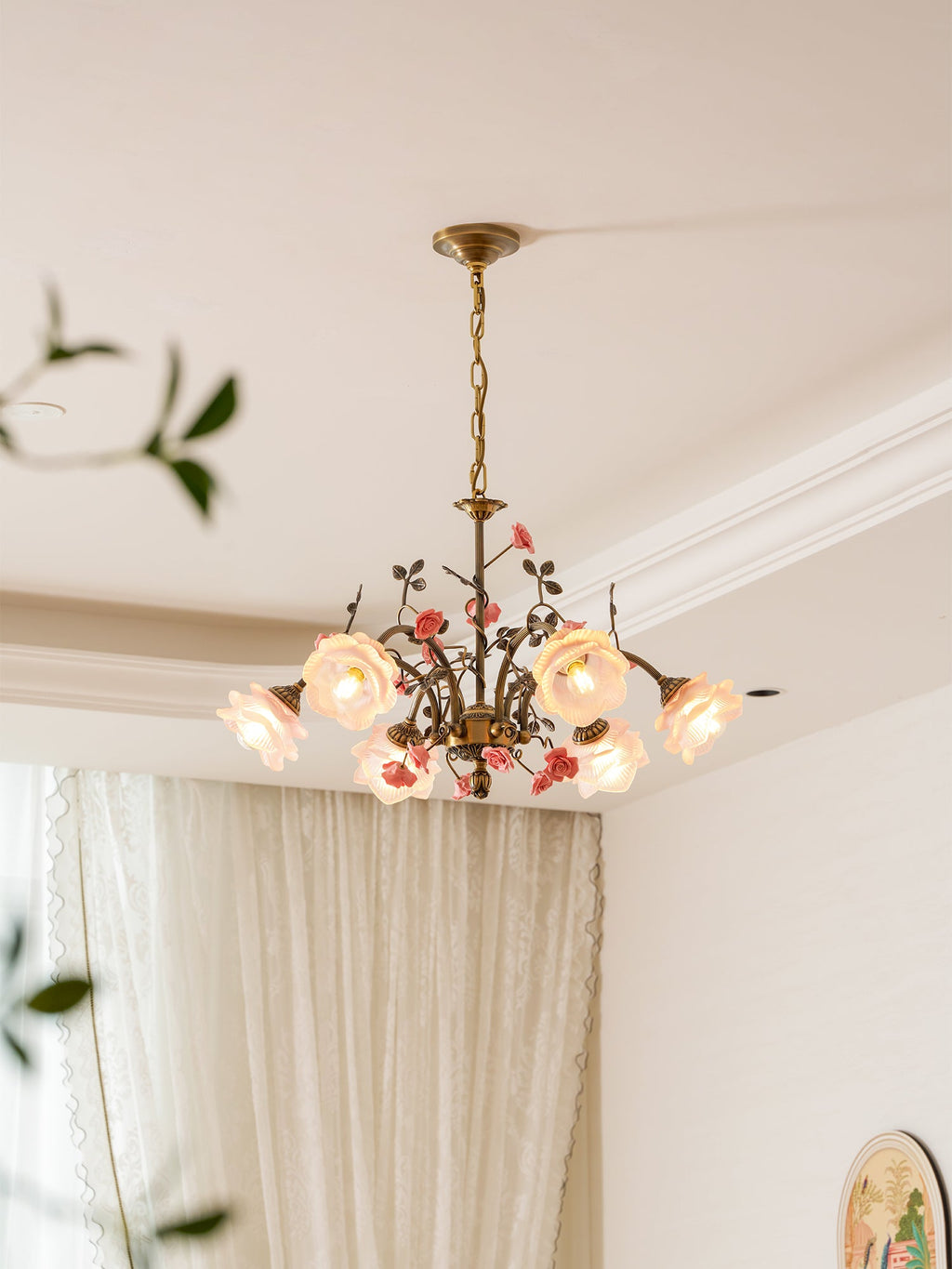 Clérachic rose chandelier lit in a room with cream curtains and cornice mouldings, with a trailing plant at the left edge.