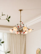 Clérachic rose chandelier lit in a room with cream curtains and cornice mouldings, with a trailing plant at the left edge.