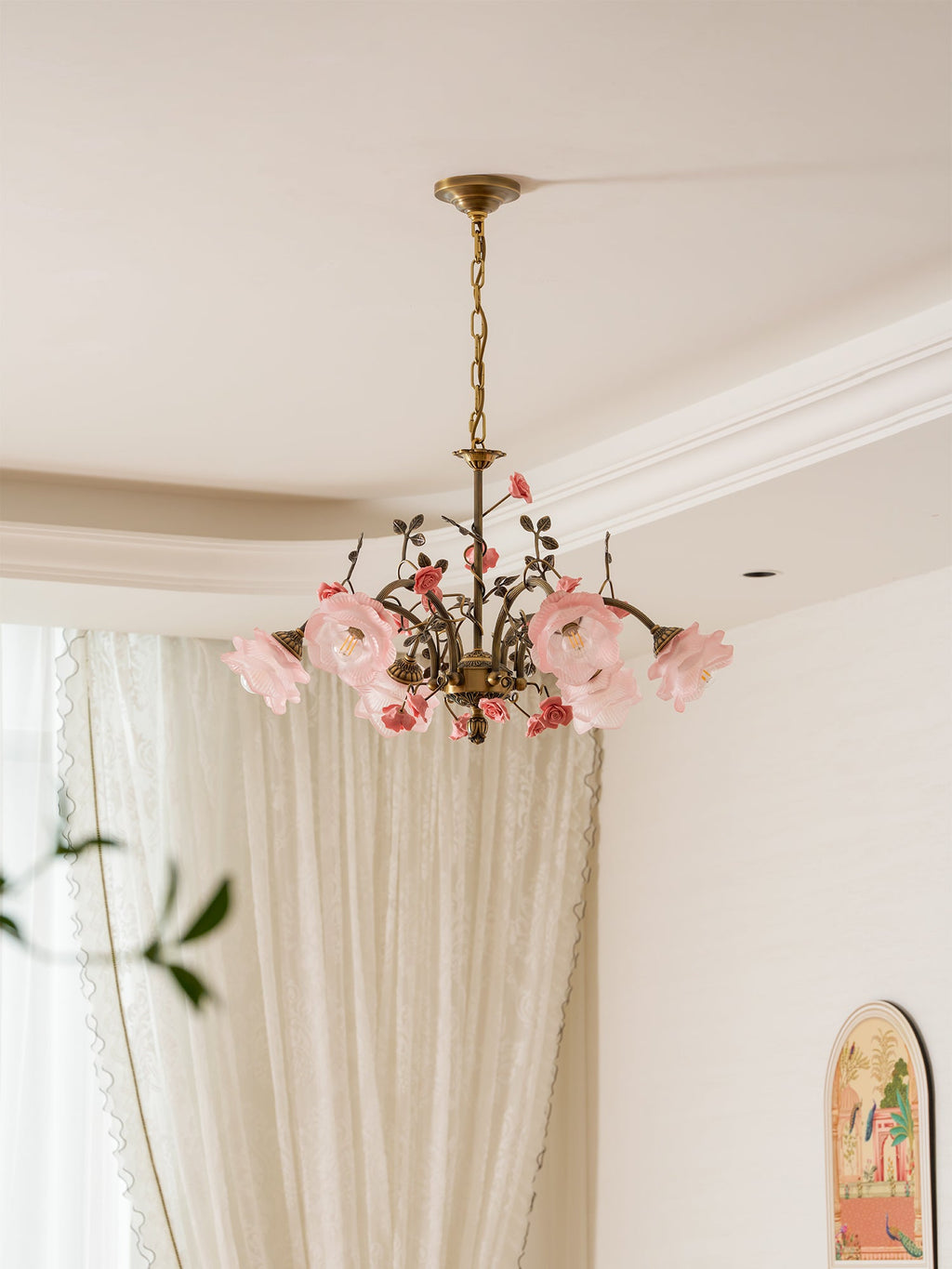 Clérachic rose chandelier seen from below with its shades unlit, pale pink against a white ceiling and cornice, in front of long curtains.