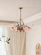 Clérachic rose chandelier seen from below with its shades unlit, pale pink against a white ceiling and cornice, in front of long curtains.