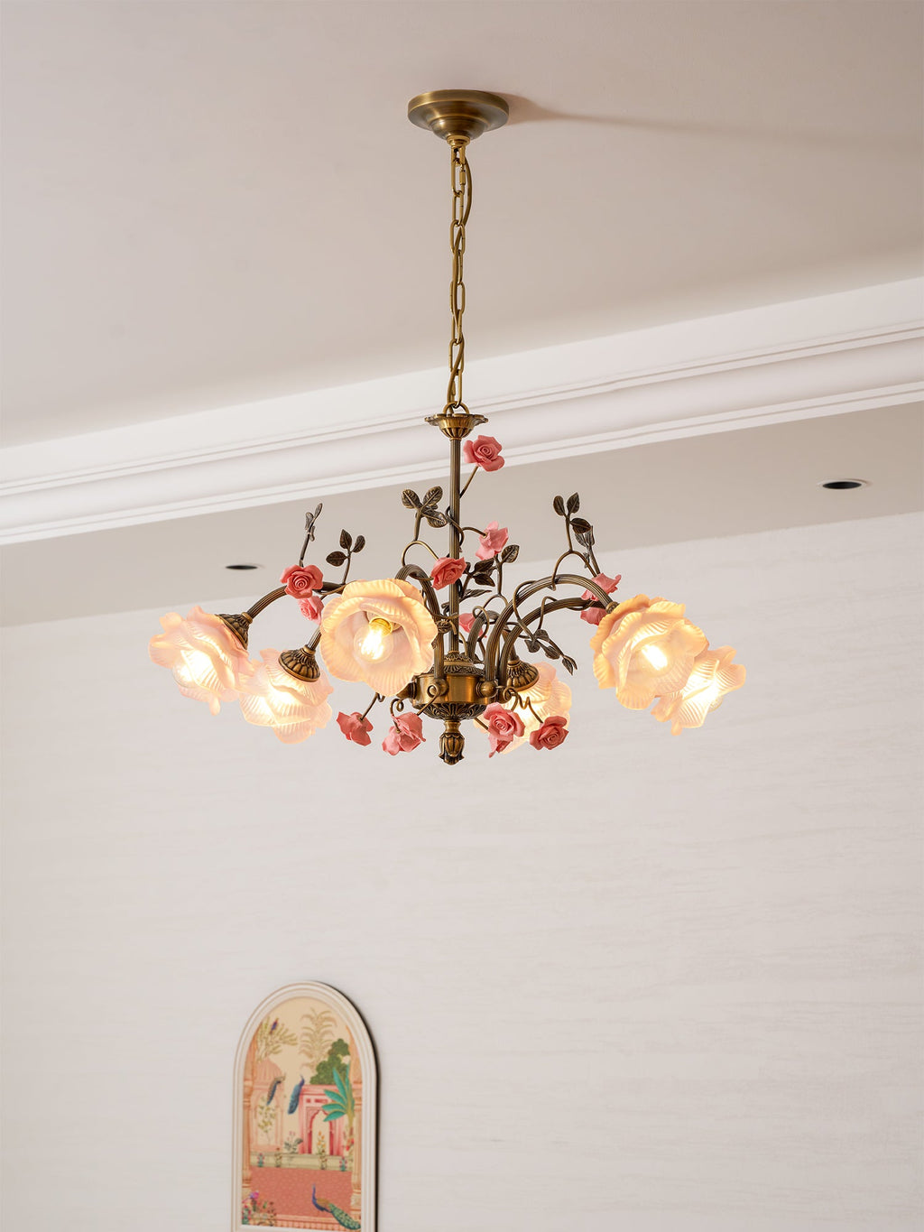 Clérachic chandelier lit from a low angle, with glass rose shades glowing beside a white cornice and a small arched picture.