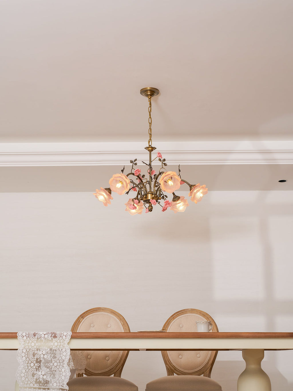 Clérachic rose chandelier lit high in a white room, above a dining table with round-backed cream chairs.