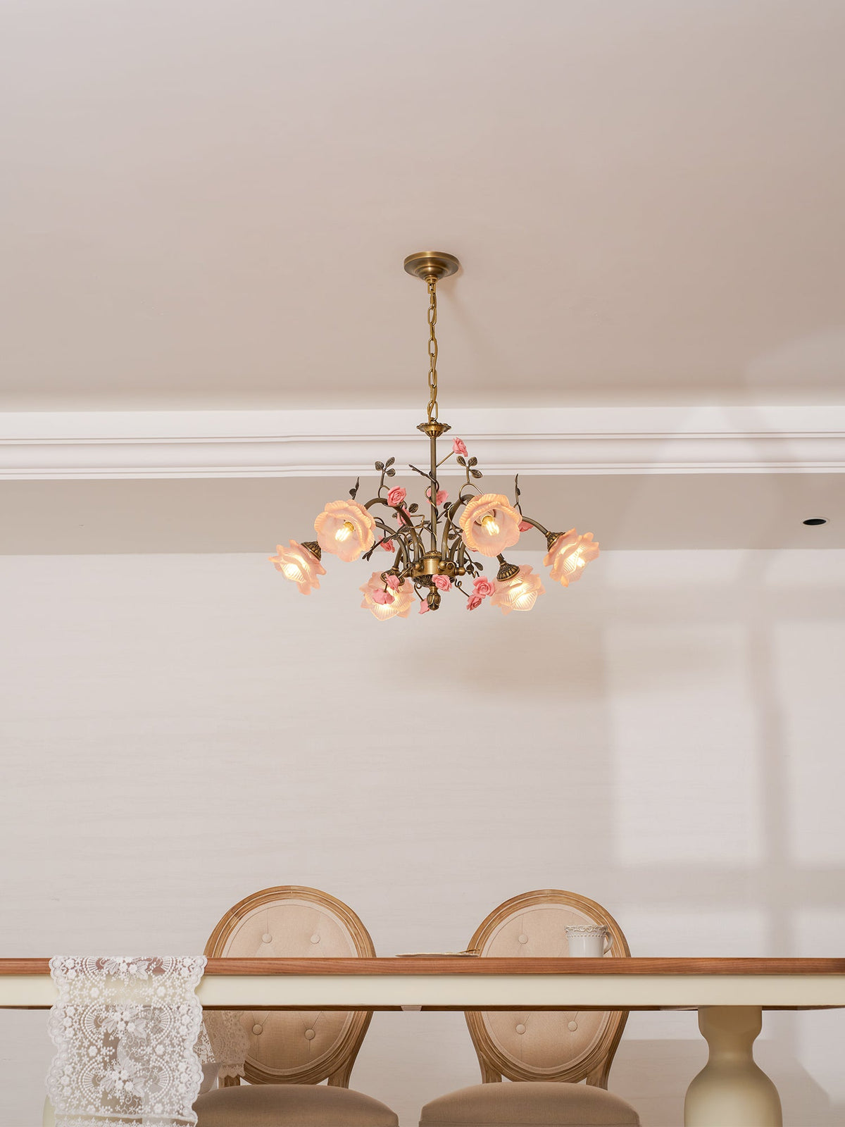 Clérachic rose chandelier lit high in a white room, above a dining table with round-backed cream chairs.