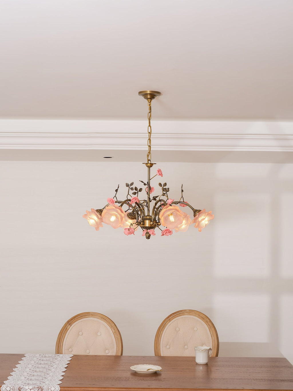 Clérachic chandelier lit with its shades glowing pink-orange, hung above a dining table with a lace runner and upholstered chairs.