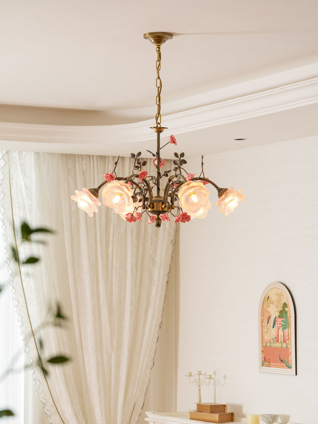 Clérachic chandelier lit in a cream room with sheer curtains, the glass roses glowing beside a plant branch and an arched picture.