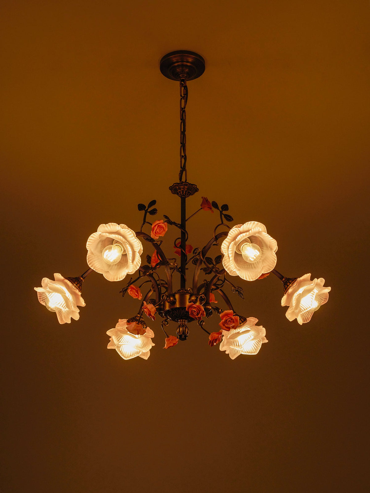 Clérachic rose chandelier glowing in a darkened room, its frosted glass roses lit amber and its ceramic roses in deep orange-pink.