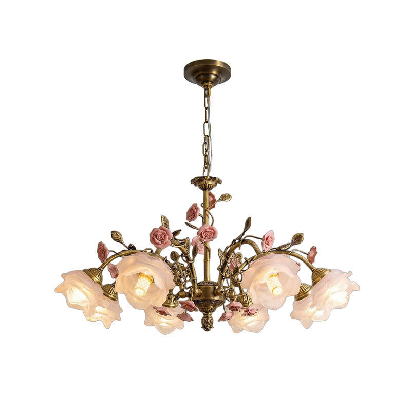 Clérachic chandelier on a white backdrop, with larger glass rose shades lit warm and pink ceramic roses between the gold arms.