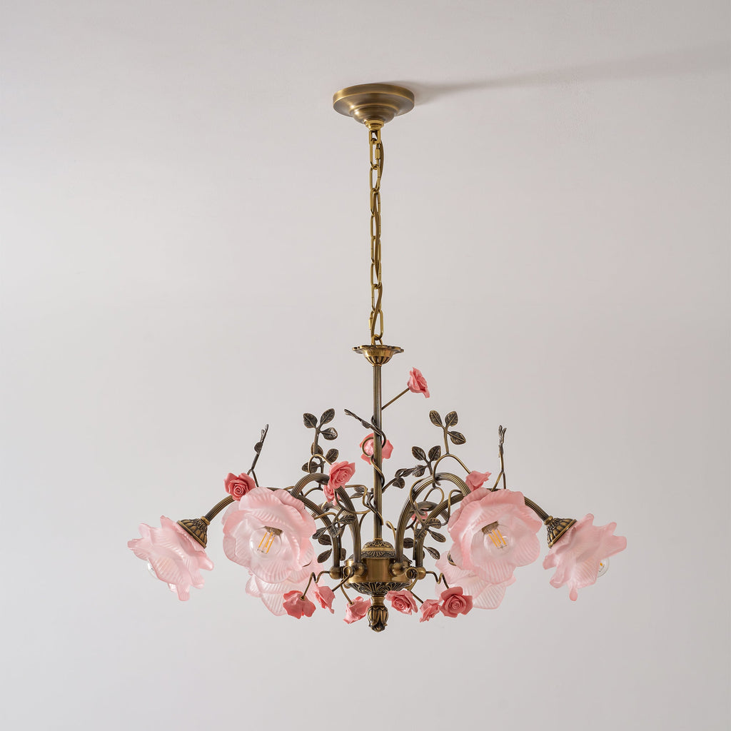 Clérachic chandelier with pink-tinted glass rose shades lit on a pale grey wall, its antique gold chain hung from a brass canopy.