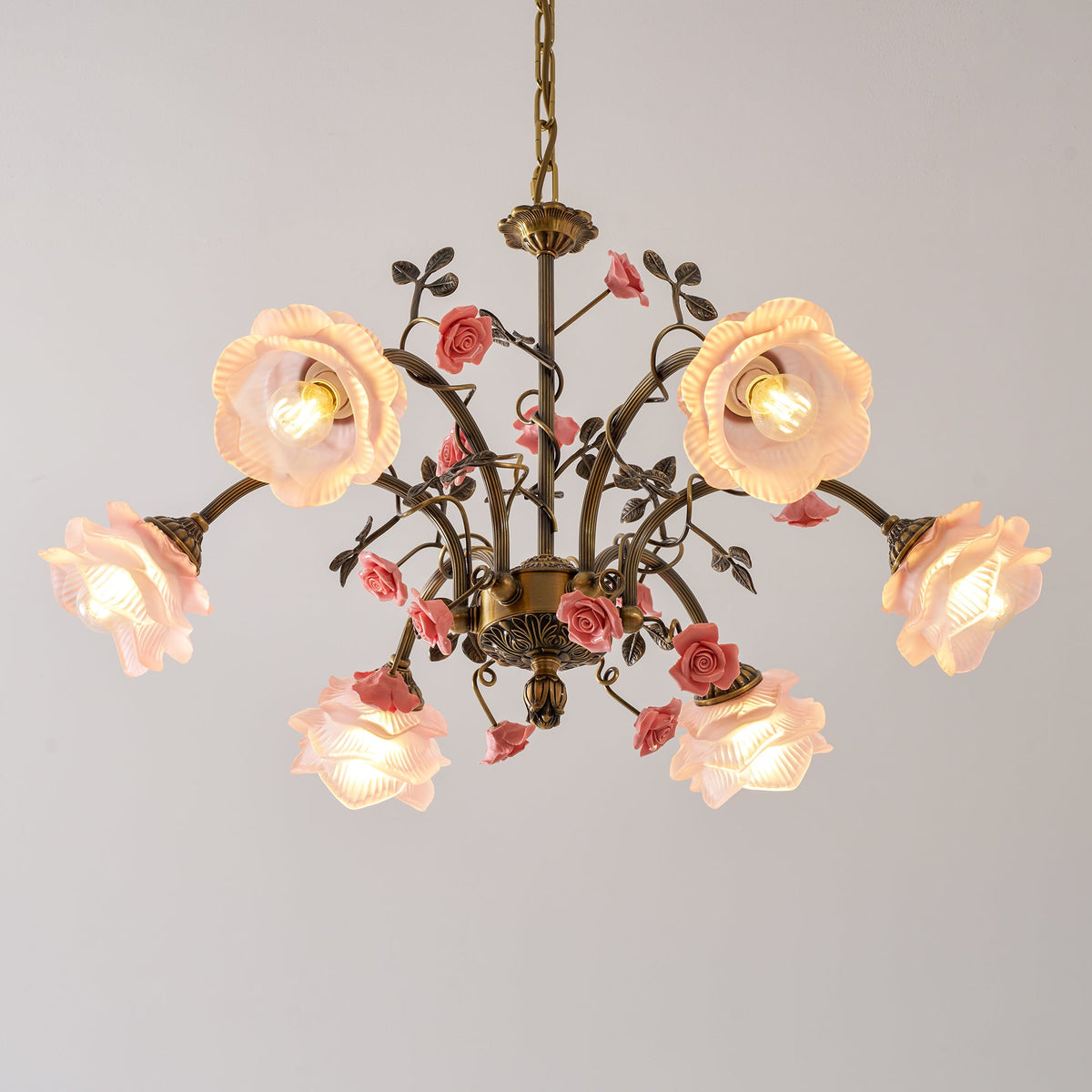 Close view of a Clérachic chandelier with lit pink frosted glass rose shades, with ceramic roses and leaves on antique gold arms.