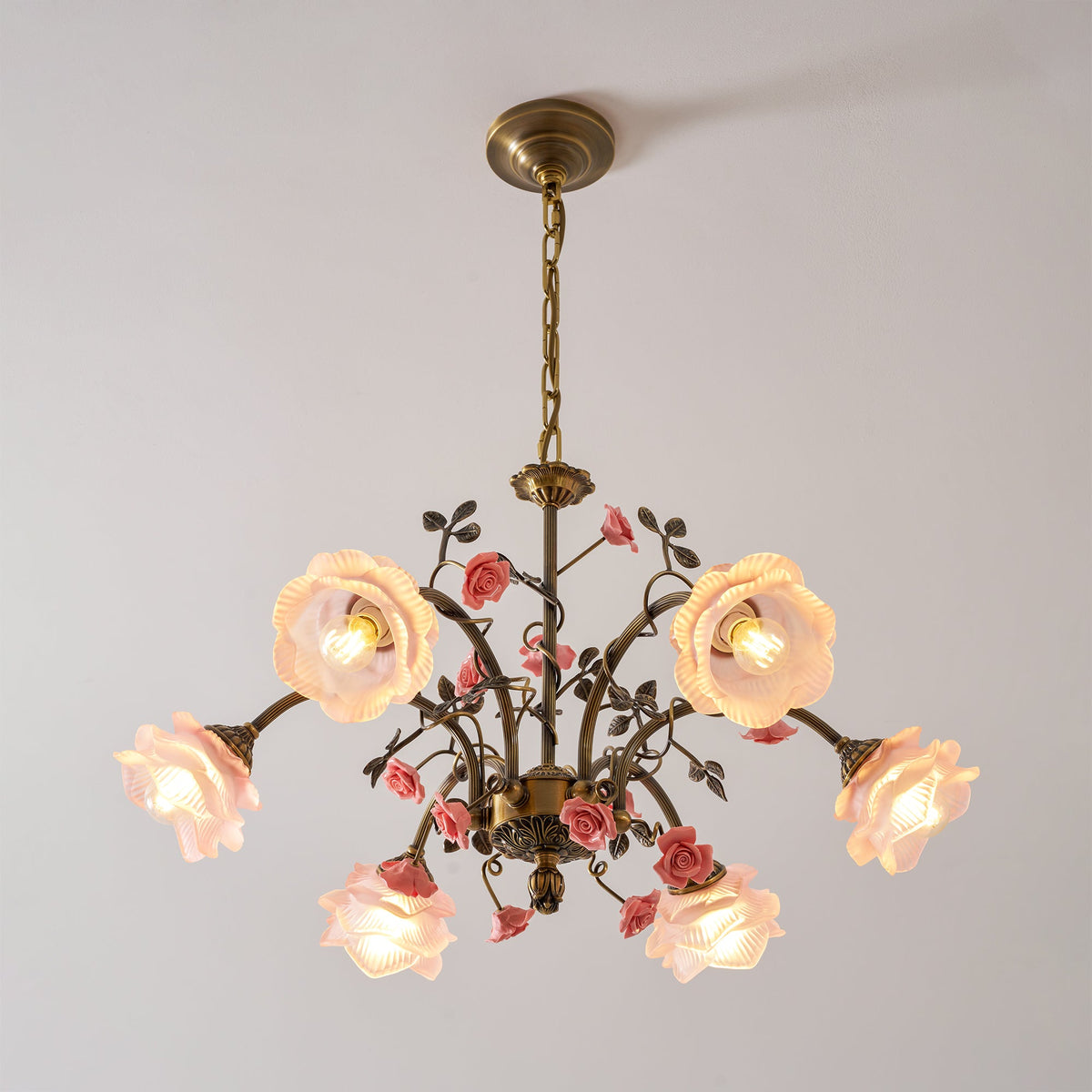 Clérachic chandelier with pink glass roses lit on a pale backdrop, seen from slightly below, with gold arms and pink ceramic roses.