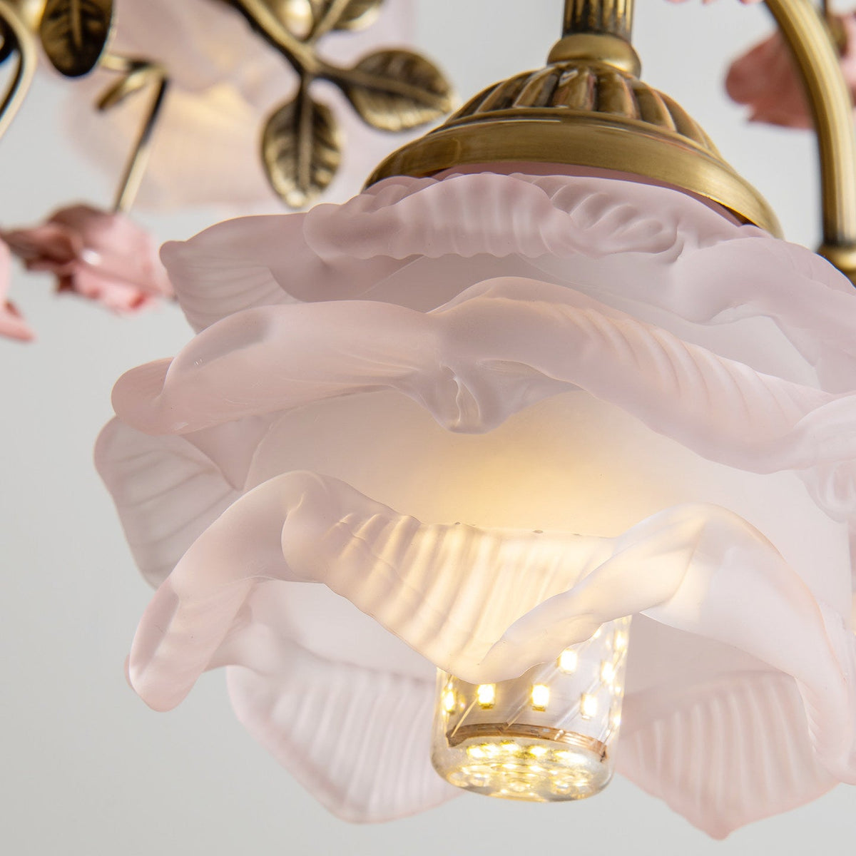 Close view of a pink frosted glass rose shade on this Clérachic chandelier, with a lit bulb inside and a gold cap and leaves above.