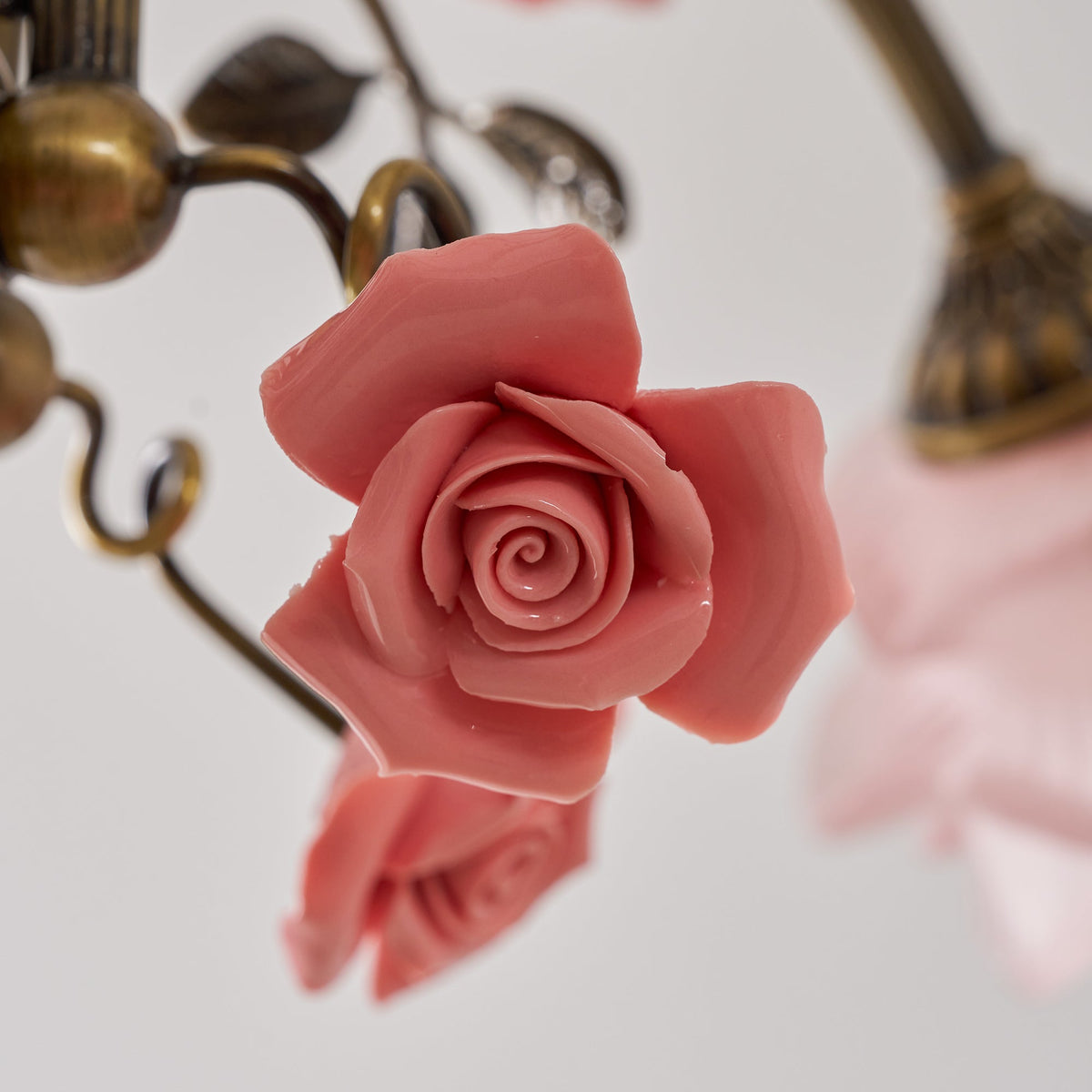 Close view of a pink ceramic rose and gold curling stems on this Clérachic chandelier, with a glass shade soft in the background.