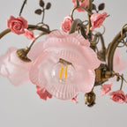 Close view of a pink glass rose shade with a filament bulb on this Clérachic chandelier, surrounded by ceramic roses and gold leaves.