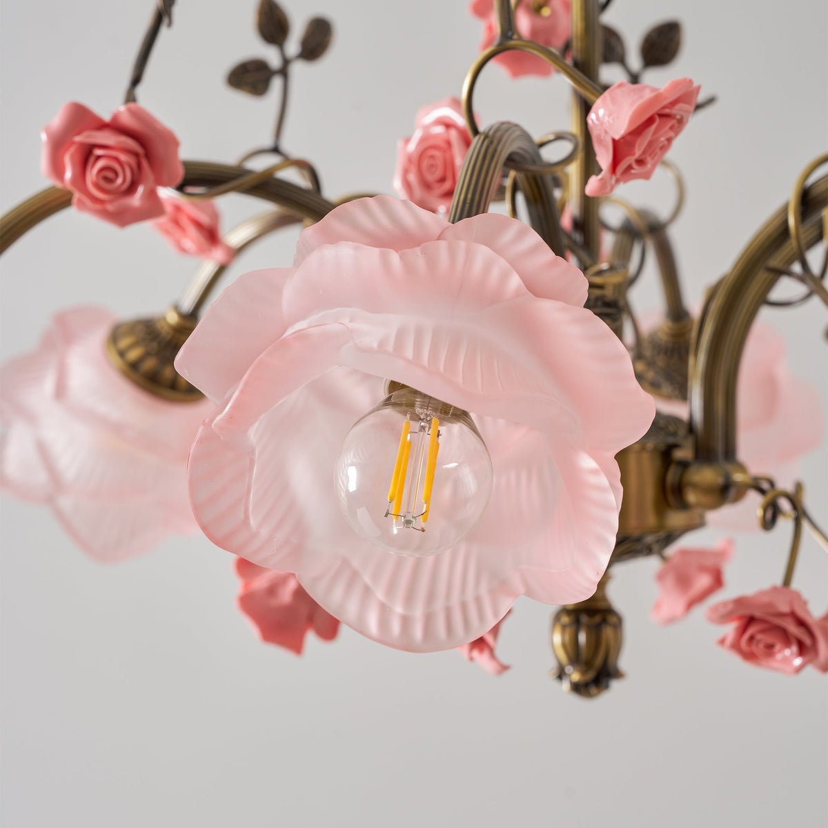 Close view of a pink glass rose shade with a filament bulb on this Clérachic chandelier, surrounded by ceramic roses and gold leaves.