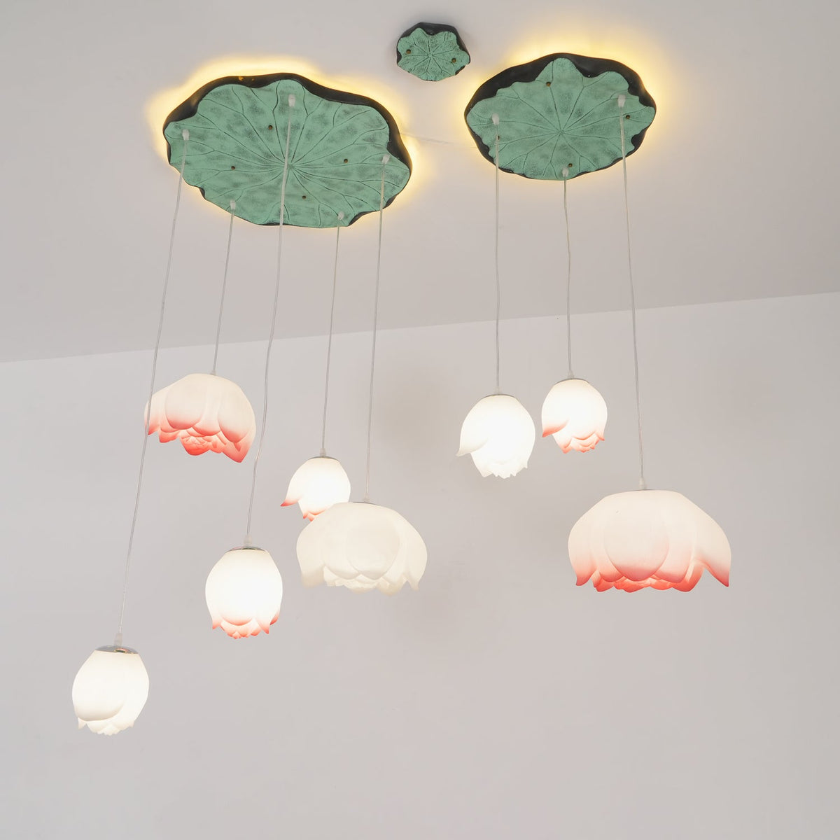 Pink-tipped and white water-lily pendants from Clérachic hang on thin cords beneath two green lily-pad canopies, against a pale wall.
