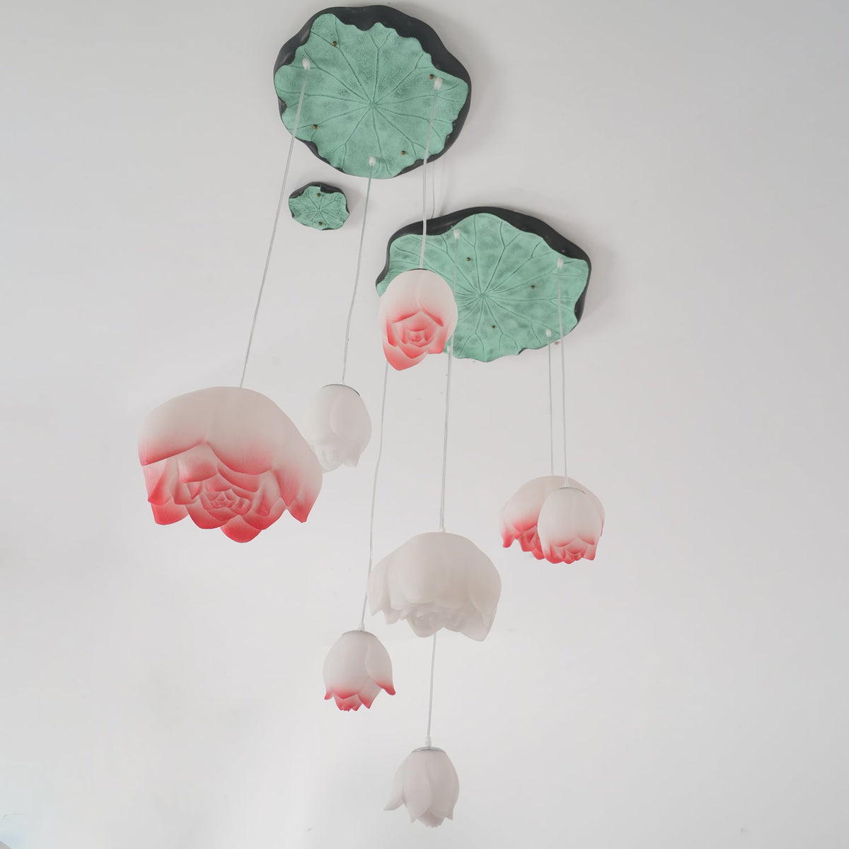 Unlit and seen from below, the Clérachic water-lily pendants show white and pink-tipped petals under green lily-pad canopies with a faint halo.