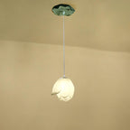 A single Clérachic water-lily pendant in a pale white bud shape hangs on a thin cord from a small green lily-pad canopy, against a warm cream wall.