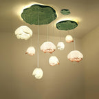Seven Clérachic water-lily pendants hang at varied heights beneath a large and a small green lily-pad canopy, glowing warm in a cream corner.