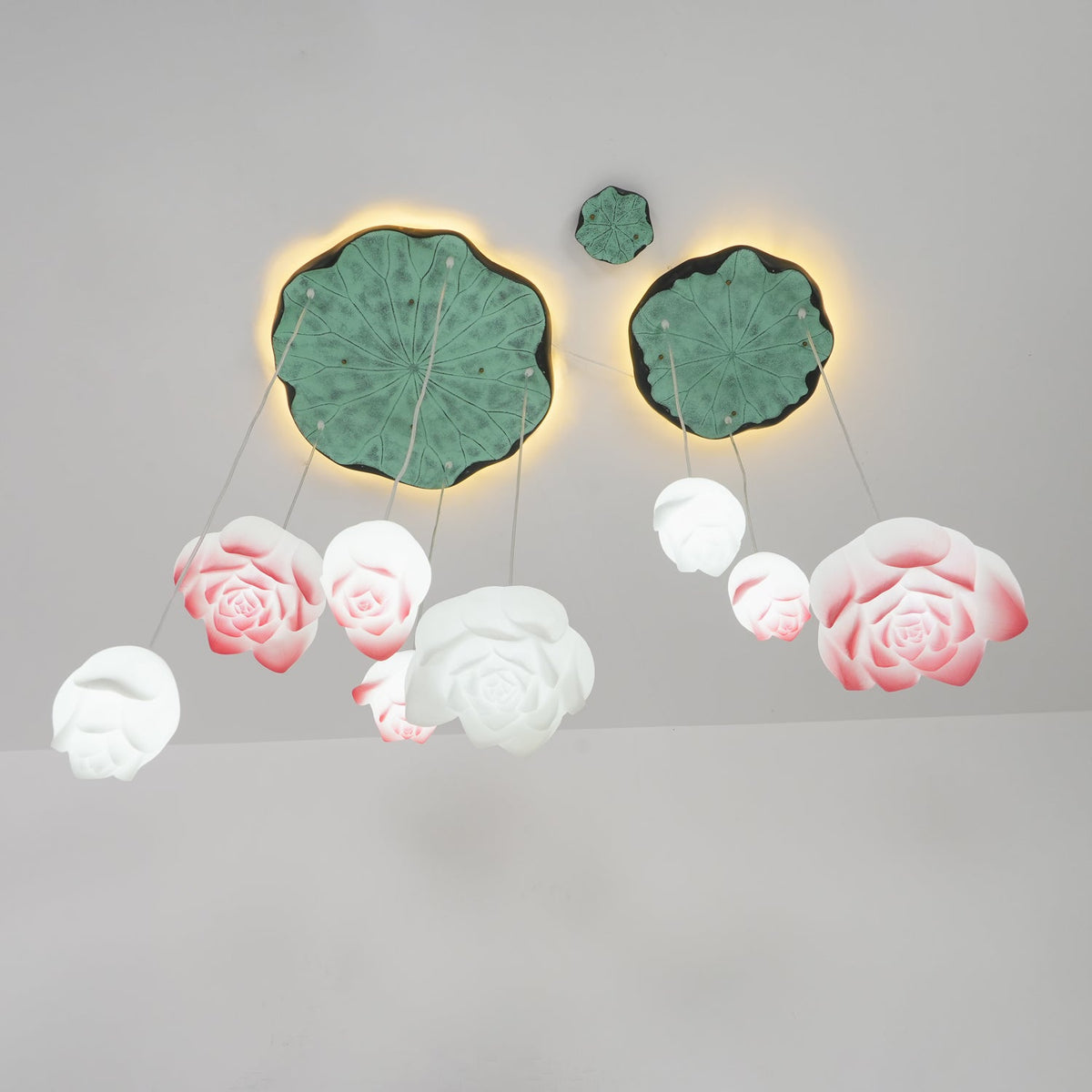 Pink-edged blooms and smaller white buds of this Clérachic water-lily chandelier, hanging in varied sizes beneath two lily-pad canopies on a grey wall.