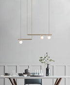 Gold Clérachic pendant with three opal globes hanging against a pale grey panelled wall, above a table set with bowls and glasses.