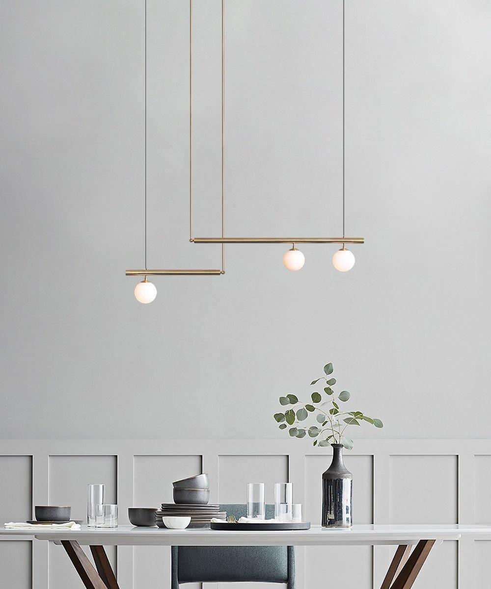 Gold Clérachic pendant with three opal globes hanging against a pale grey panelled wall, above a table set with bowls and glasses.