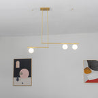 Three-globe gold Clérachic pendant on a white wall, with offset bars hanging from a ceiling plate, beside two abstract prints.