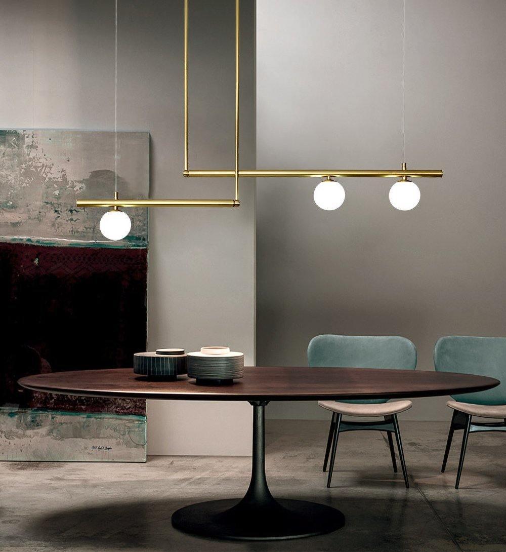 Clérachic gold pendant with offset bars and three glowing globes above a round walnut table, with teal chairs and a large painting.