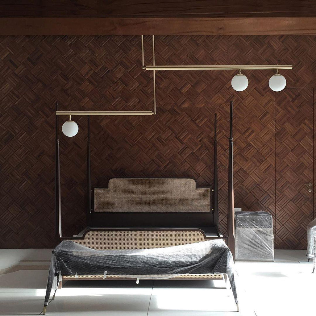 Clérachic gold pendant with three globes hanging in front of a herringbone timber wall, over a bed with a rattan headboard.