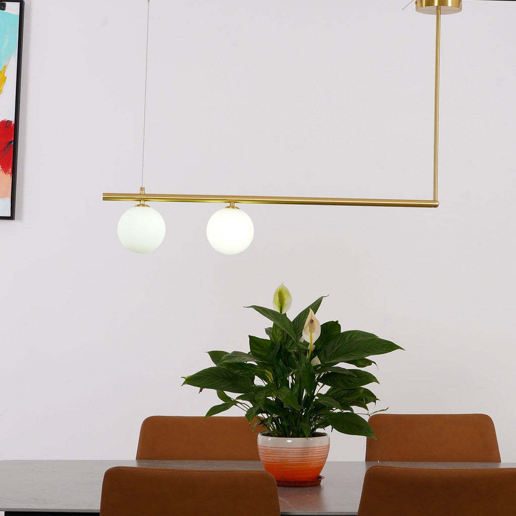 Clérachic two-globe gold pendant bar over a dining table, with a peace lily in a pot and tan leather chairs below.