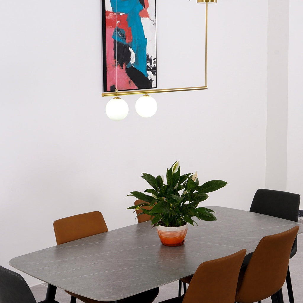 Close view of a two-globe Clérachic gold pendant over a grey-topped dining table, with a plant and a pink and blue print.