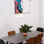 Close view of a two-globe Clérachic gold pendant over a grey-topped dining table, with a plant and a pink and blue print.
