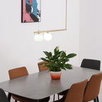 Two opal globes on a gold Clérachic pendant bar, hung over a dining table with a peace lily and tan chairs.