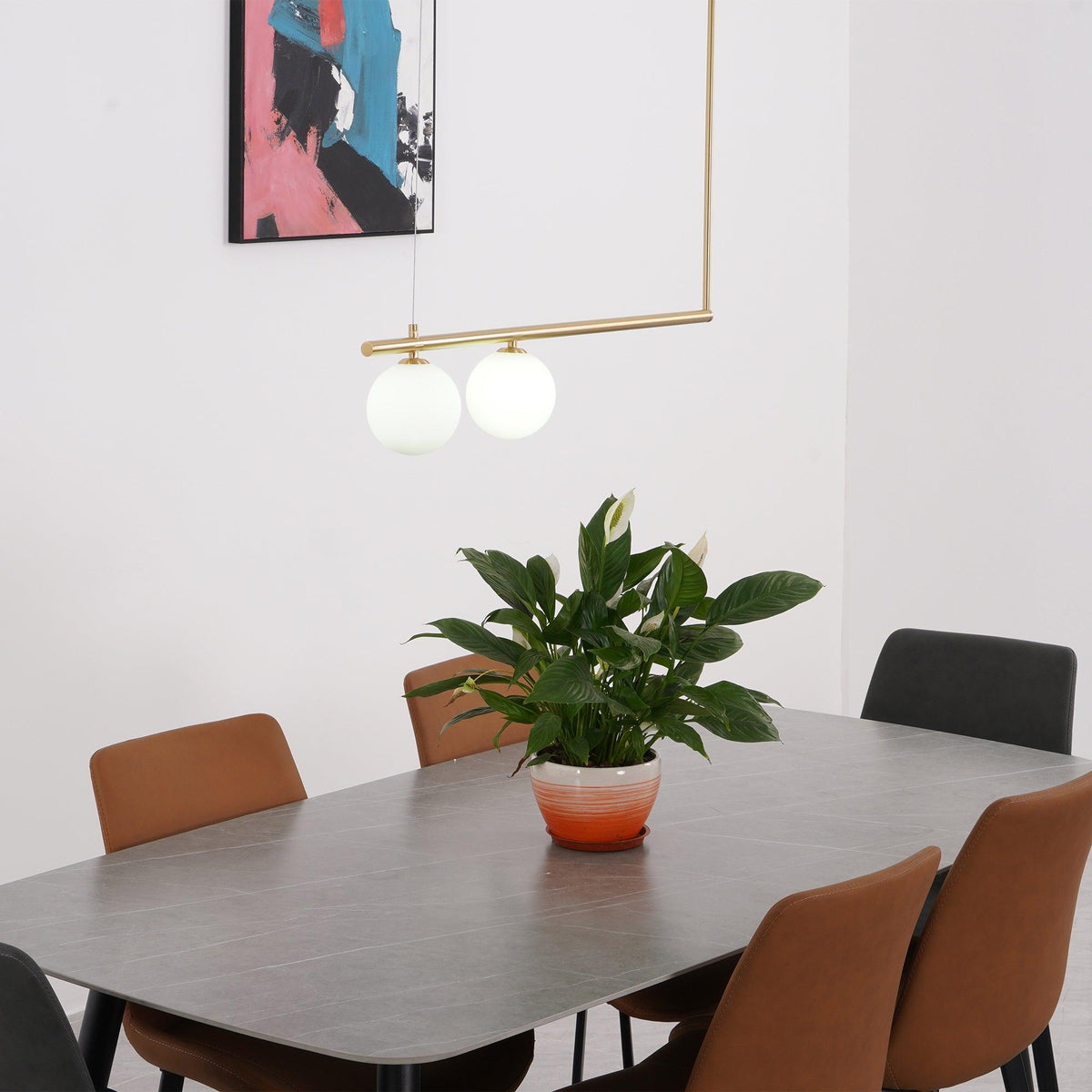 Two opal globes on a gold Clérachic pendant bar, hung over a dining table with a peace lily and tan chairs.
