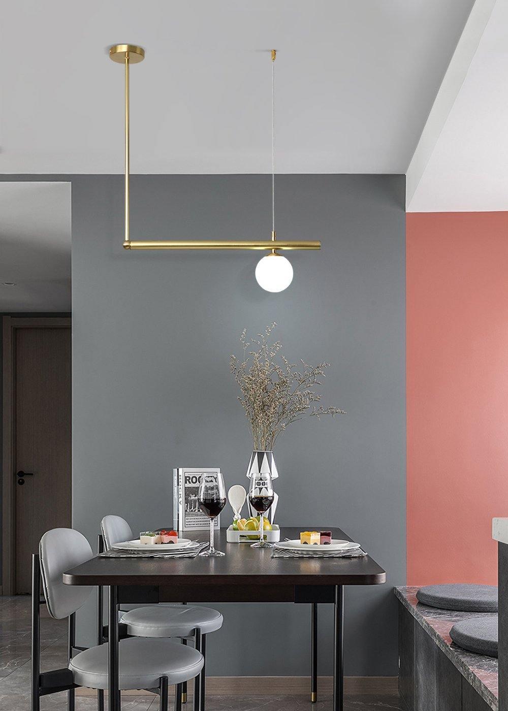 Single-globe Clérachic pendant on a gold rod above a dark dining table, with a grey wall and a coral panel behind.