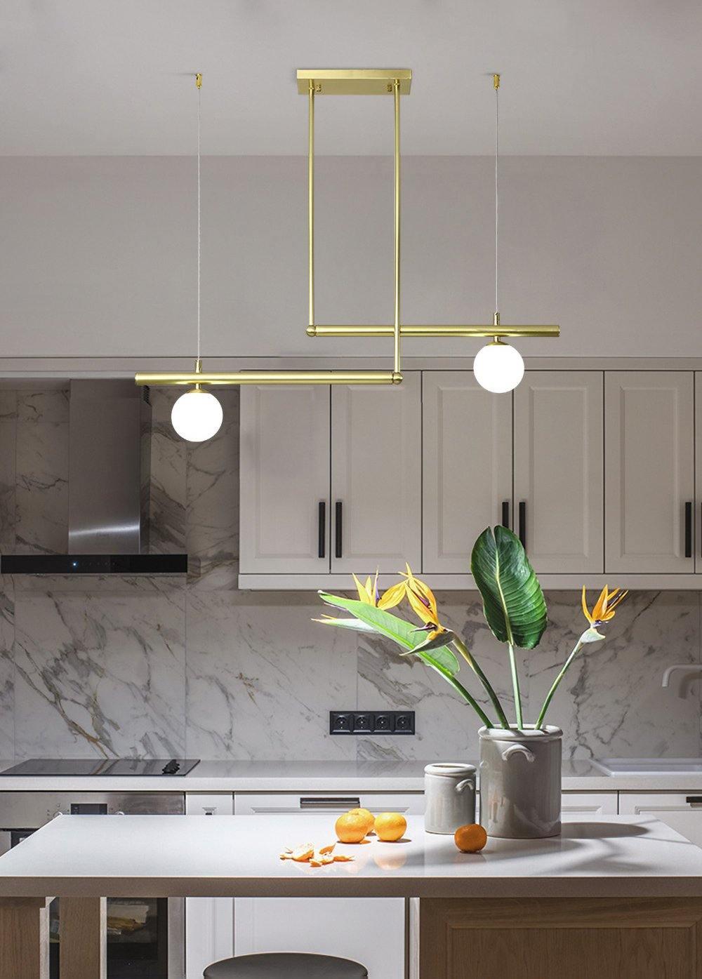 Two-globe Clérachic gold pendant above a white kitchen island, with a vase of leaves and fruit, and marble splashback behind.