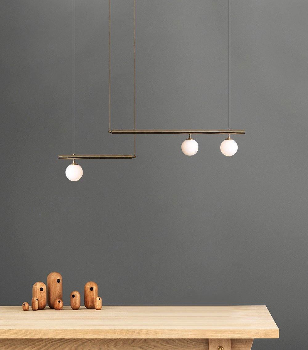 Clérachic gold pendant with three opal globes against a dark grey wall, above a pale wood table with small wooden figures.