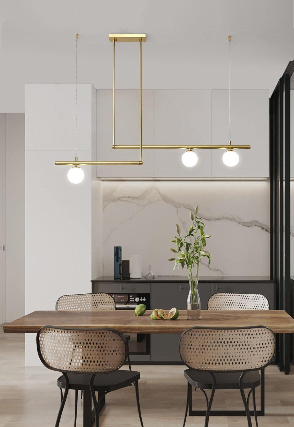 Clérachic gold pendant with three globes hung above a wooden dining table with woven chairs, with a marble wall behind.