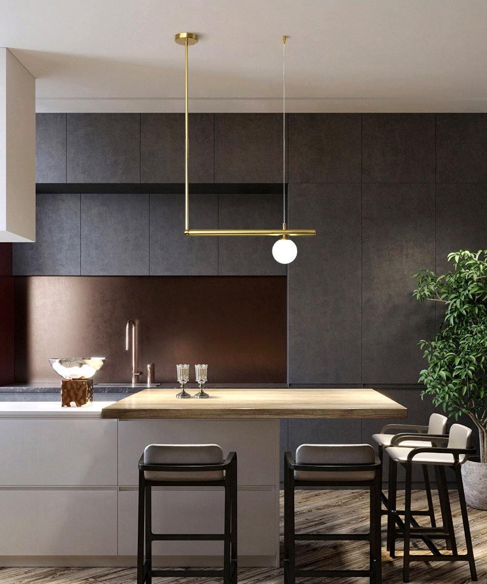Single-globe Clérachic pendant on a gold rod above a kitchen island in a dark-toned kitchen, with black bar stools.