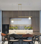 Clérachic gold pendant with two offset bars and two glowing globes in a dark timber kitchen, above a dining table.