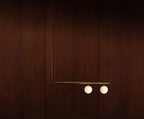 Two-globe Clérachic pendant with a long rod on a dark mahogany wall, its gold bar and opal globes softly lit.