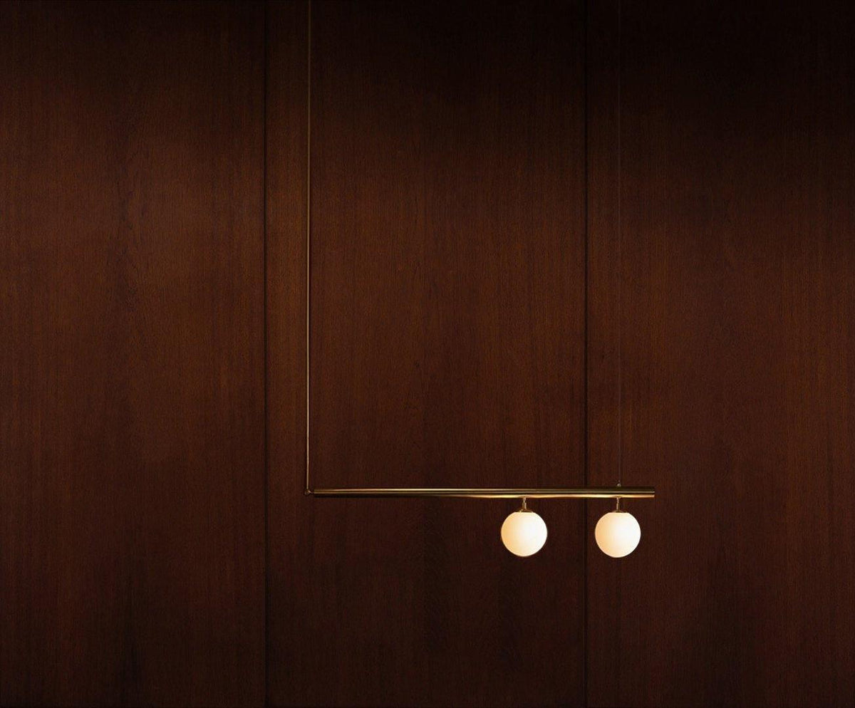 Two-globe Clérachic pendant with a long rod on a dark mahogany wall, its gold bar and opal globes softly lit.