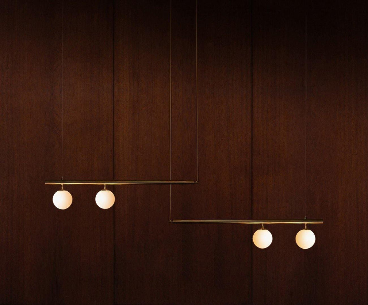 Clérachic pendant with two bars carrying four lit opal globes, hanging against a dark mahogany wall.